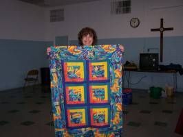 Person holding a colorful patchwork quilt in a hall with a cross on the wall.