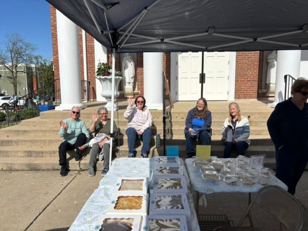 People seated at a table under a dark canopy with trays of food outdoors on a sunny day