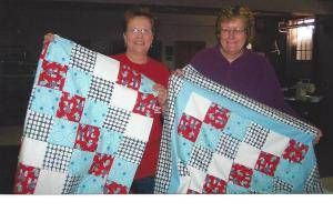 Two people holding a blue, red, and white patchwork quilt indoors