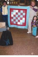 Three people hold a red-and-white quilt in a room, with a black bag in the foreground.