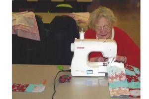 Woman sewing at a table with a white sewing machine and colorful fabric pieces nearby.