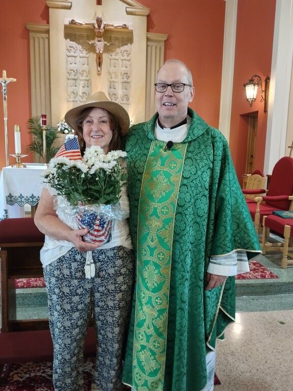 Woman holding flowers stands beside a priest in a church sanctuary, both smiling.