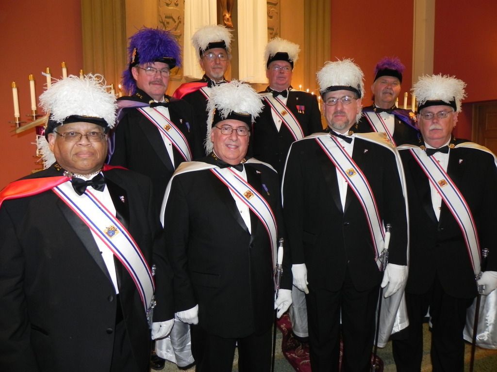 Group of uniformed performers in black suits, white gloves, and tall white hats posing indoors.