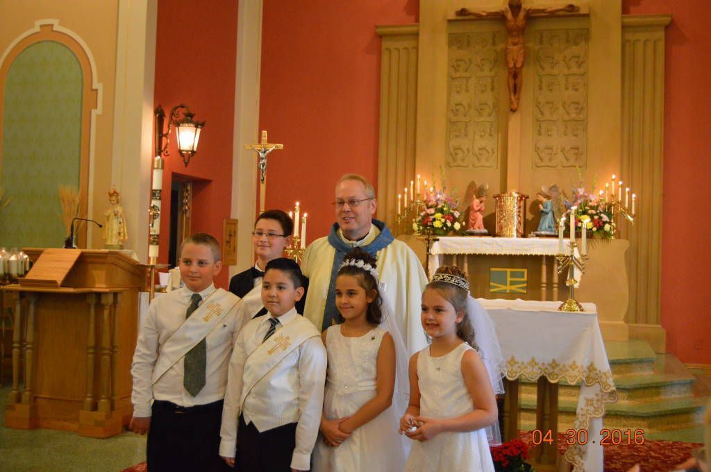 Children in white outfits stand in a church before the altar with a priest behind them.
