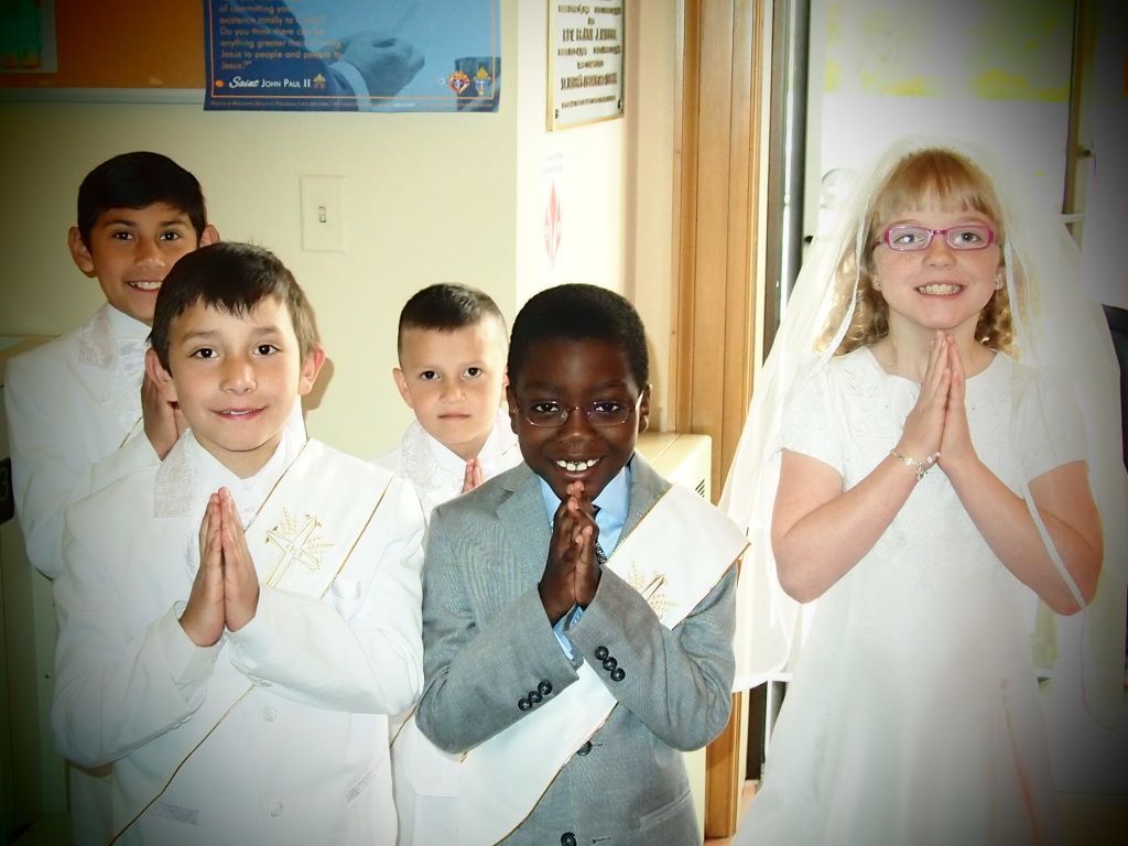 Children in white clothing stand with hands together in a classroom, smiling at the camera.