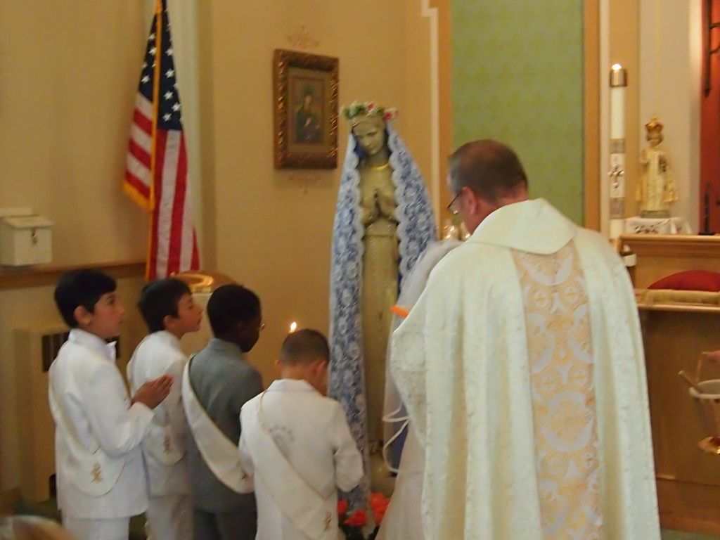 Priest and children gathered at a Catholic altar beside a statue of Mary and a U.S. flag