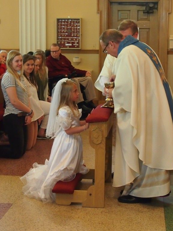 Child kneels at church altar while a priest gives communion, with seated congregation nearby.