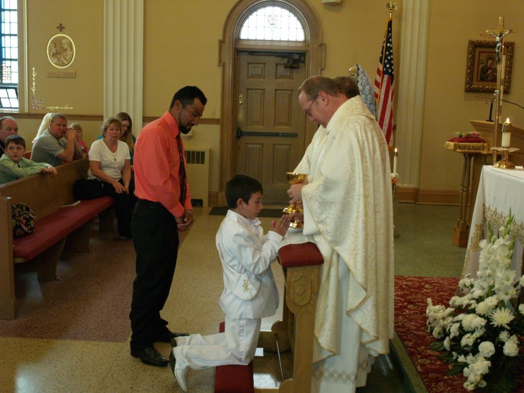 Boy kneels to receive communion from a priest in a church, with seated attendees in the background.