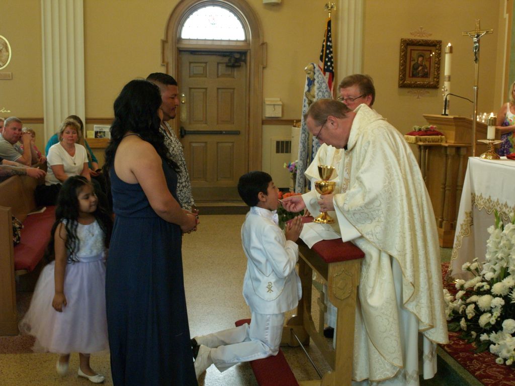 Priest giving communion to kneeling boy in white suit inside a church, with family nearby