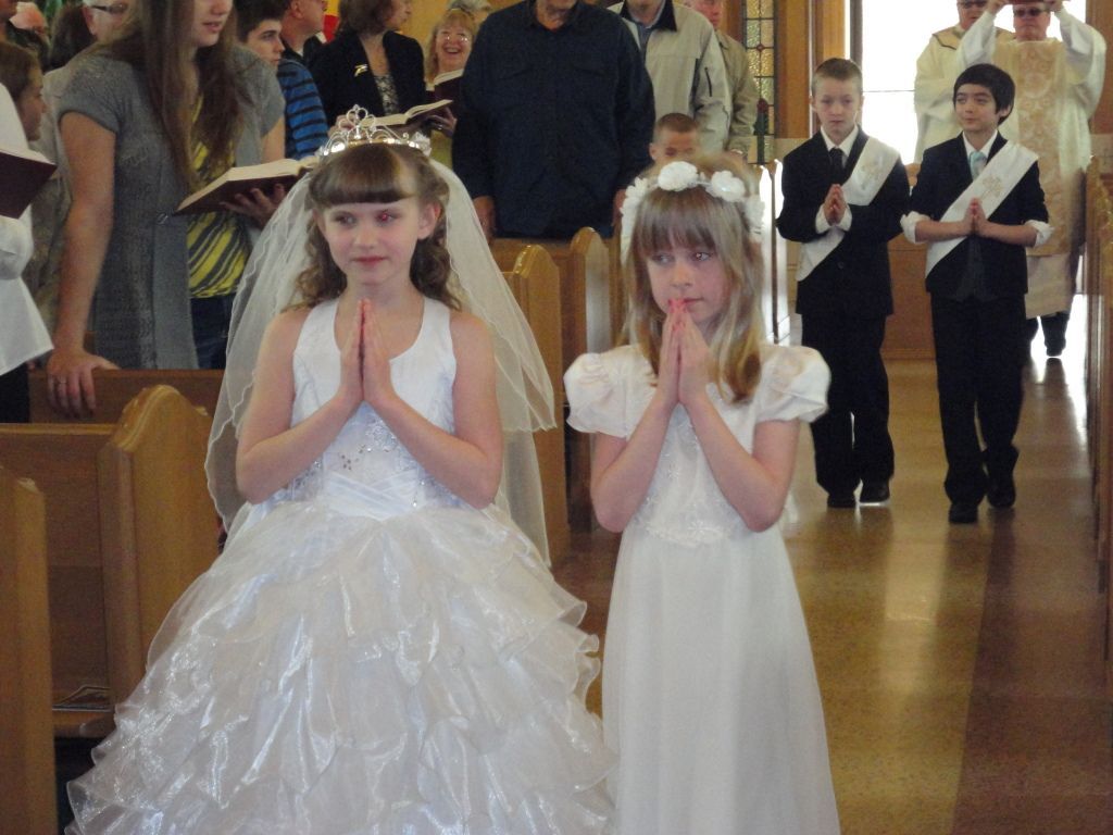 Two children in white dresses with flower crowns pose in a church aisle during a ceremony.