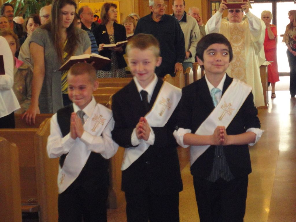 Three boys in dress clothes stand indoors holding certificates during a ceremony.