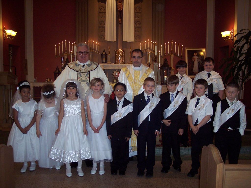 Group posed in a church before an altar, with two priests and children in white outfits and sashes.