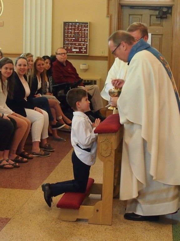 Boy kneels at church altar with priest during a service, while seated congregation watches nearby