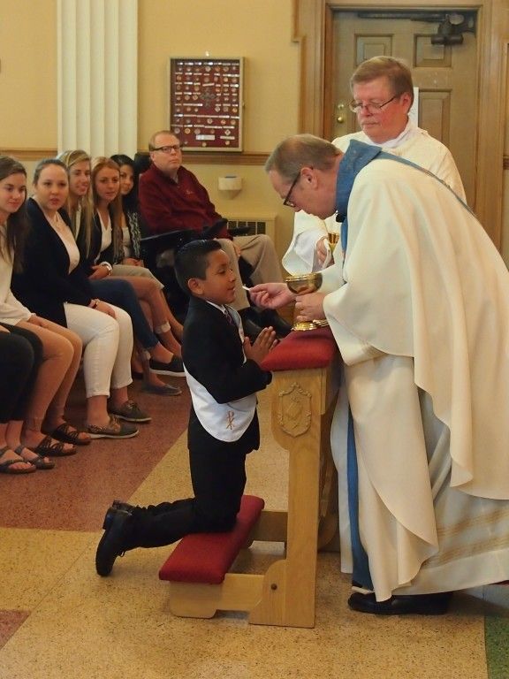 Child kneeling to receive Communion from a priest in a church, with seated congregants nearby