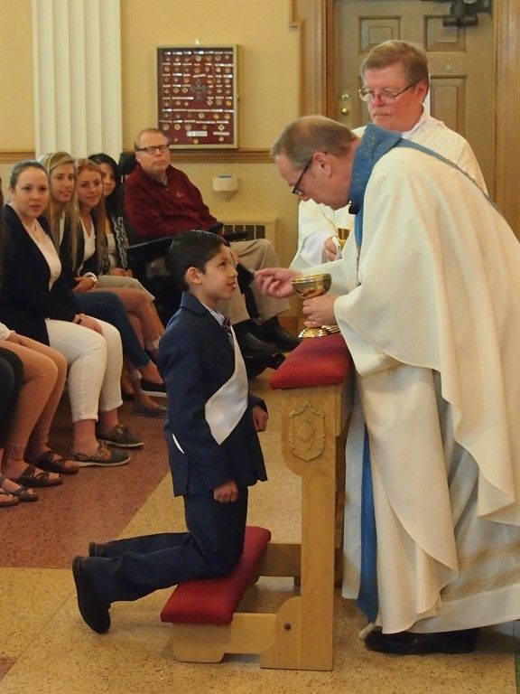 Boy kneels to receive communion from a priest in a church, with seated congregants nearby.