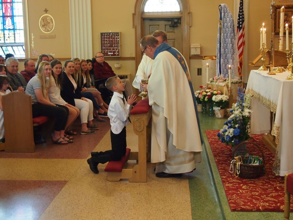 Boy kneeling before a priest during a church ceremony, with seated guests watching nearby.
