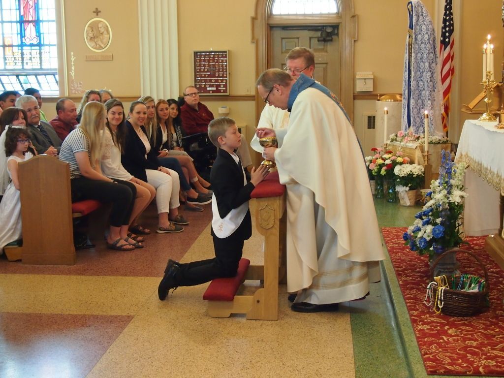 Boy kneels before priest at church altar during a religious ceremony, with seated congregation nearby