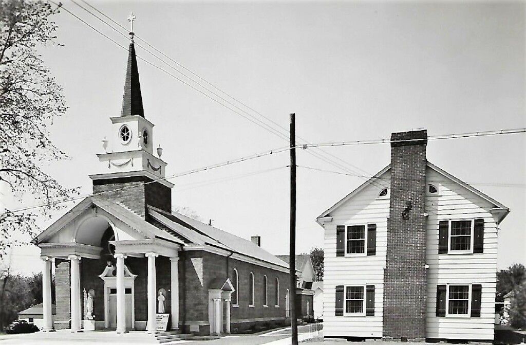 Small church with tall steeple beside a two-story white house, viewed in black and white.