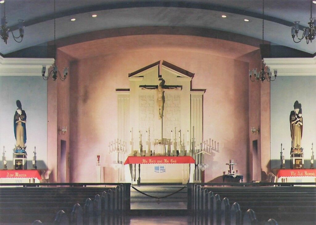 Church altar with cross and candles, flanked by statues, viewed from pews in a dim sanctuary