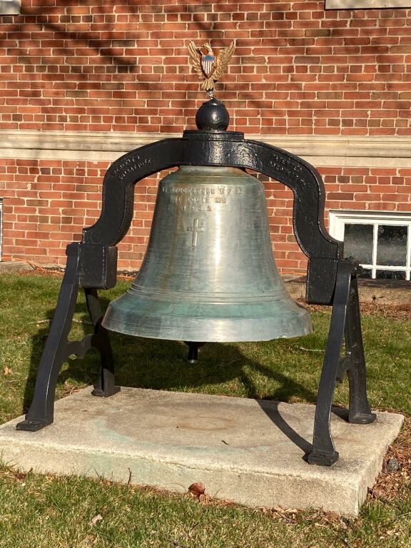 Large bronze bell on a black frame outdoors against a brick building.