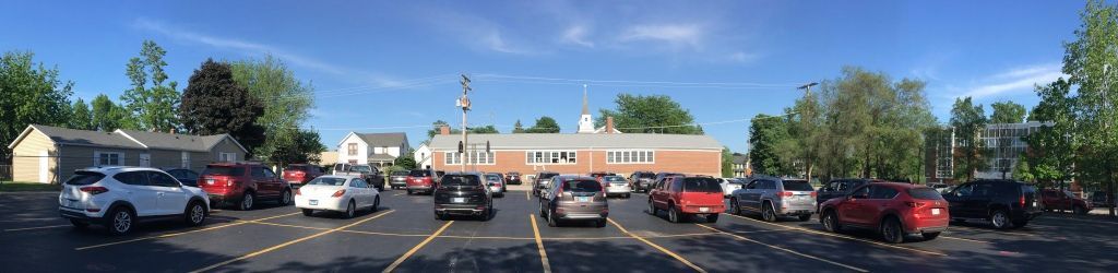 Parking lot with several cars under a bright blue sky, near a low building and trees