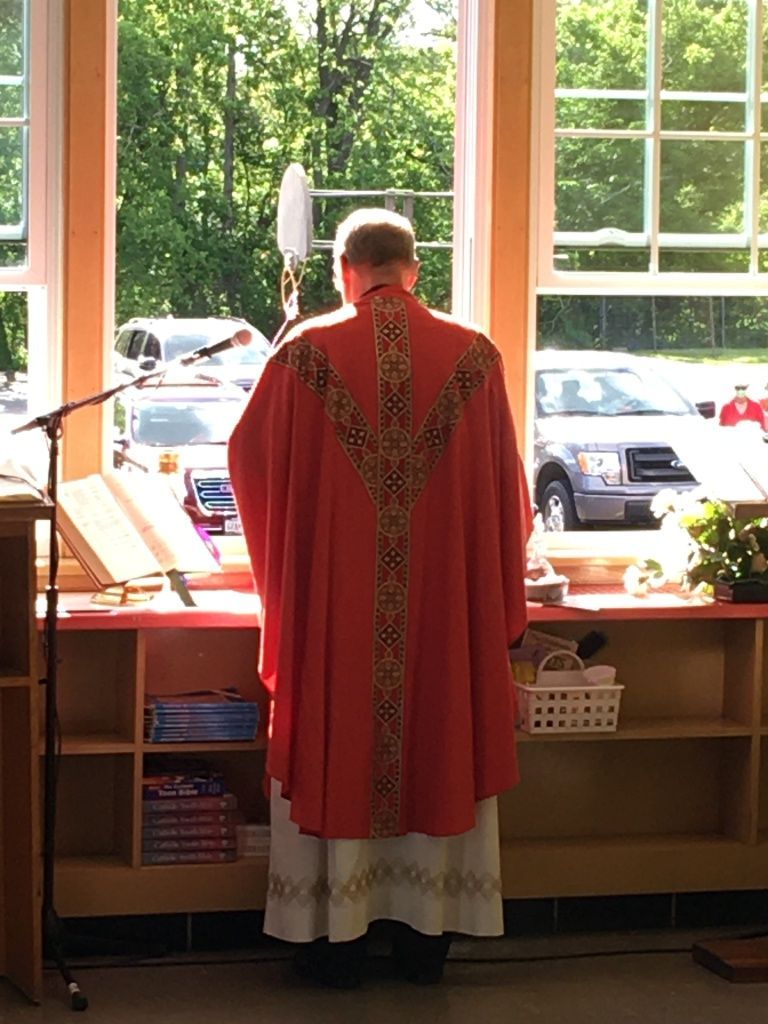 Person in red vestments stands at a windowed altar, facing outside in a church.