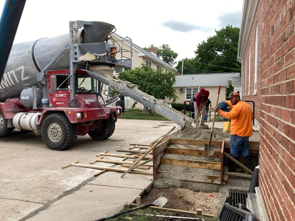 Construction workers pouring concrete from a mixer truck into a basement area beside a brick building.