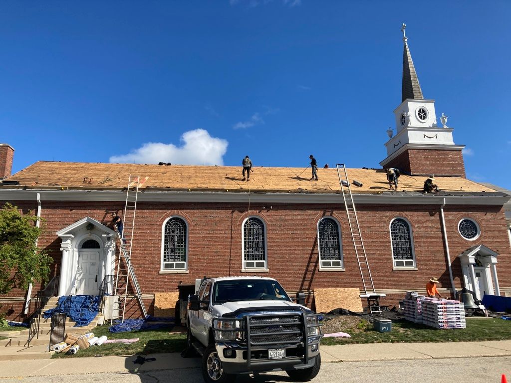 Workers on a brick church roof under repair, with a truck and church steeple visible.