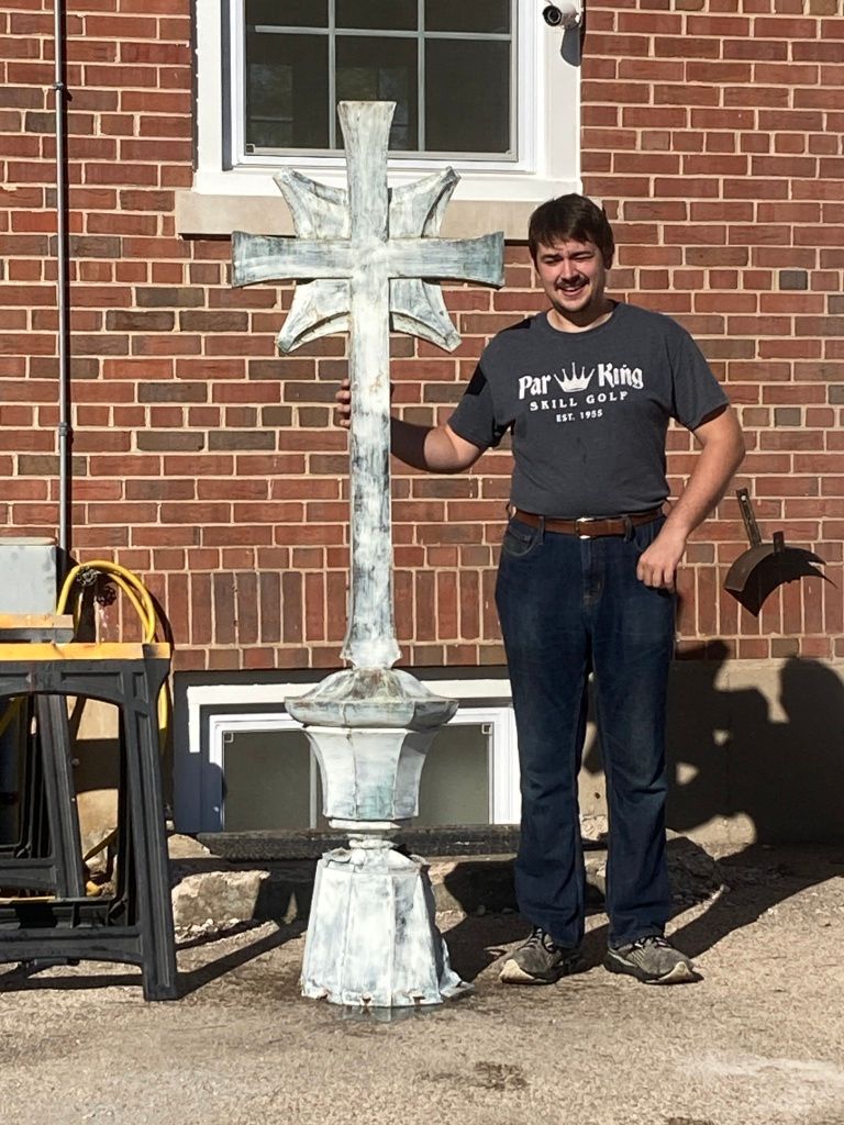 Man standing beside a large white cross sculpture in front of a brick building