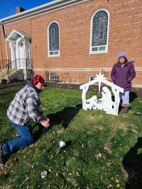 Two people by a small white nativity display on a grassy lawn beside a brick church.