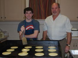 Two people in a kitchen prepare pancakes on a griddle, one holding a spatula beside several cooking pancakes.