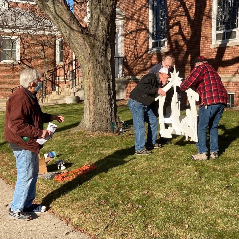 Three people assembling a white outdoor decoration on a lawn beside a brick building.