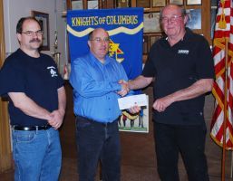 Three men at a ceremony; one presents an envelope before a Knights of Columbus banner and flag.