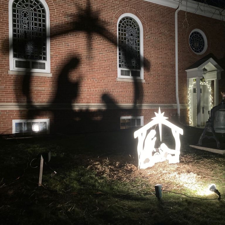 Nighttime churchyard with lit nativity scene and large shadows on a brick wall.
