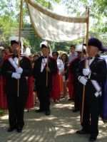 Three people in black robes and hats stand outdoors under a canopy, holding staffs.
