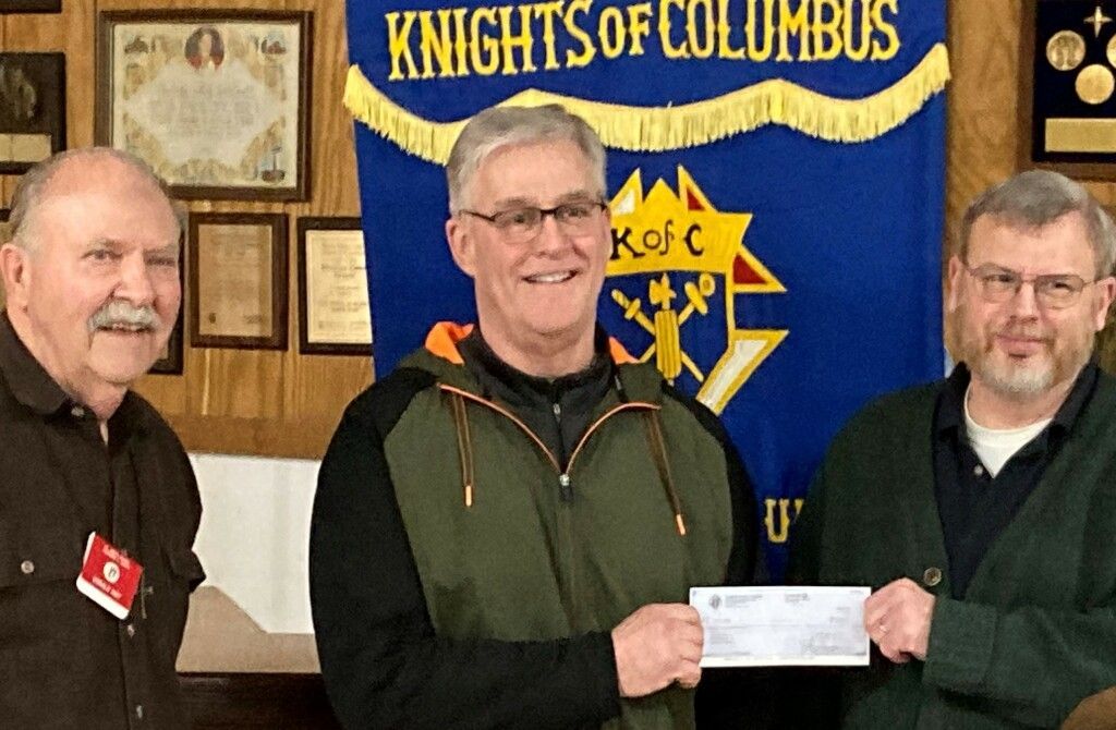 Three men stand before a Knights of Columbus banner while the center man holds a check.