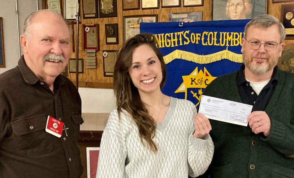 Three people posing indoors before a Knights of Columbus banner, holding a white certificate.