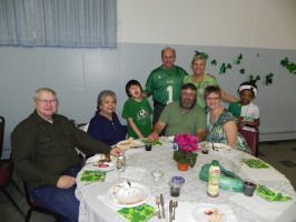 Group seated around a table in a room, with a green-clad person standing behind them and decorations on the wall.