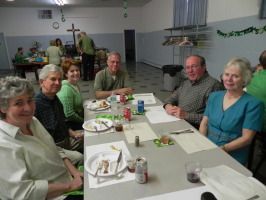 People seated at a long table in a bright hall, sharing a meal with plates, drinks, and a cross in the background