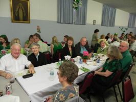 People seated at long tables in a community hall, chatting and smiling during a gathering