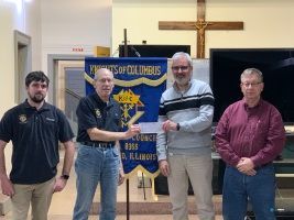 Four men stand indoors before a banner and large cross, holding a certificate and plaque.