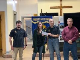 Four people standing indoors near a banner and cross, one holding a check.