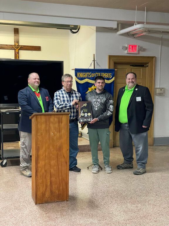 Four men stand onstage by a podium, holding a plaque in a community hall.