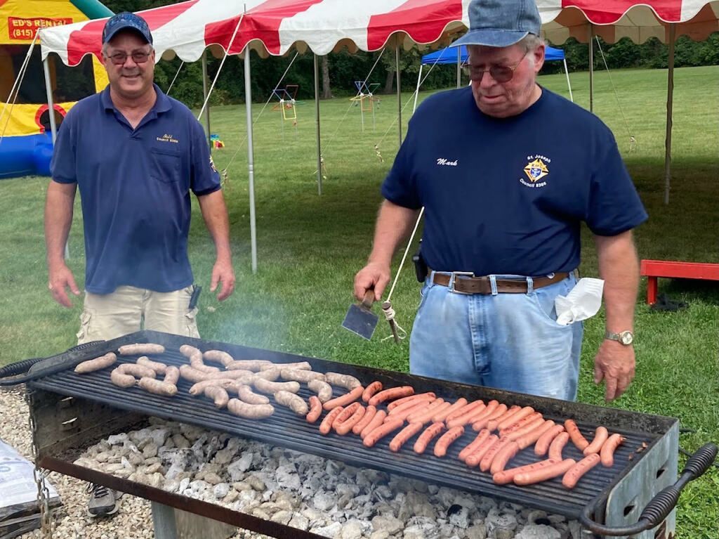 Two men grilling sausages under a red-and-white tent at an outdoor event
