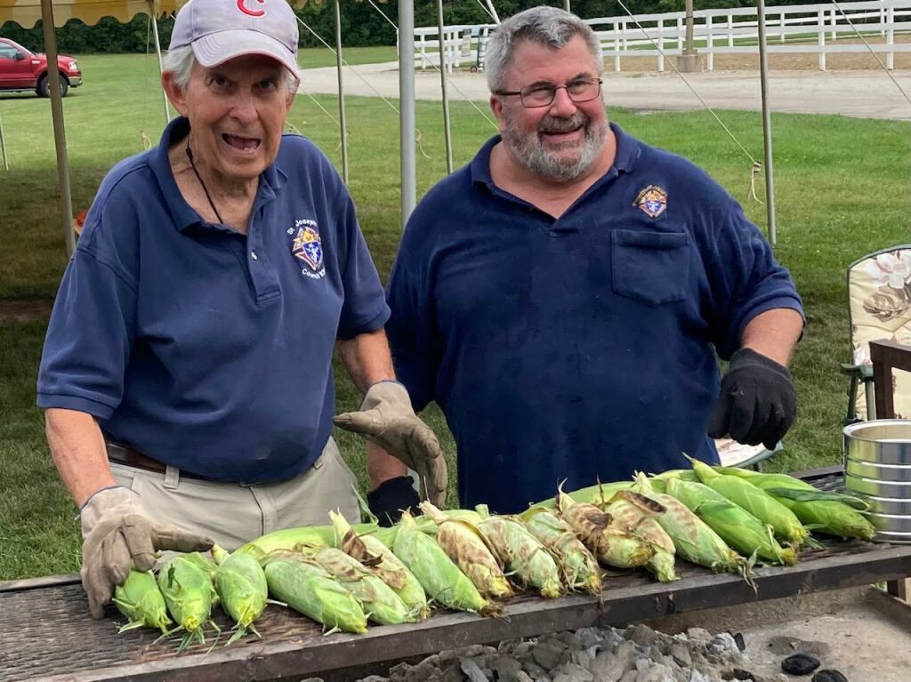 Two men in navy shirts display a row of harvested corn on a table outdoors.