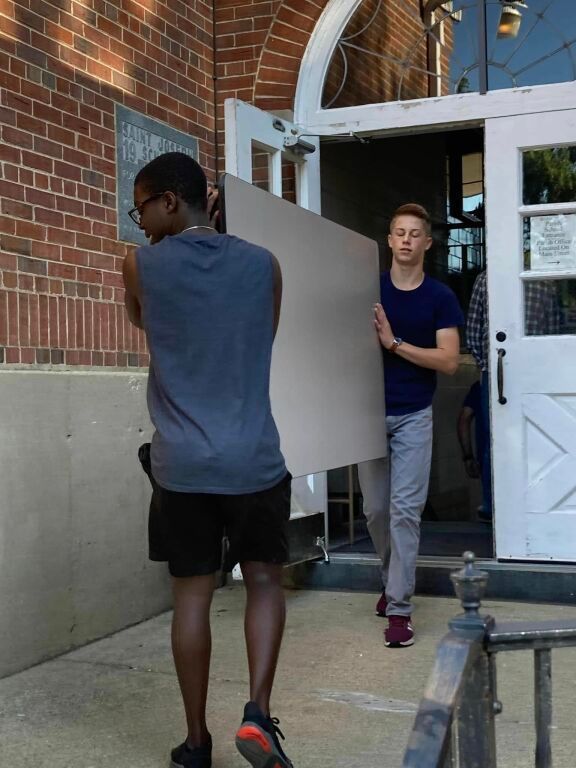 Two people carrying a large gray panel through a brick building entrance