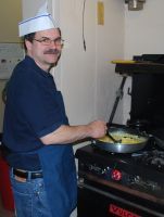 Man in a paper chef hat cooking at a stovetop, stirring food in a pan in a kitchen.