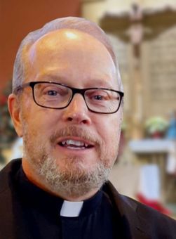 Portrait of a smiling clergy member wearing glasses and a black shirt with a white collar in a softly blurred indoor setting