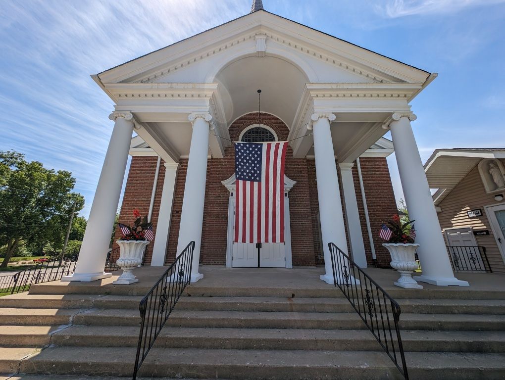 Brick church entrance with white columns, stairs, and a large American flag hanging over the doors.