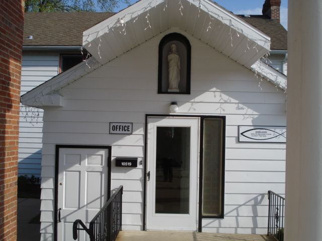 Small white office building entrance with a glass door, ramp, and religious statue above the door.